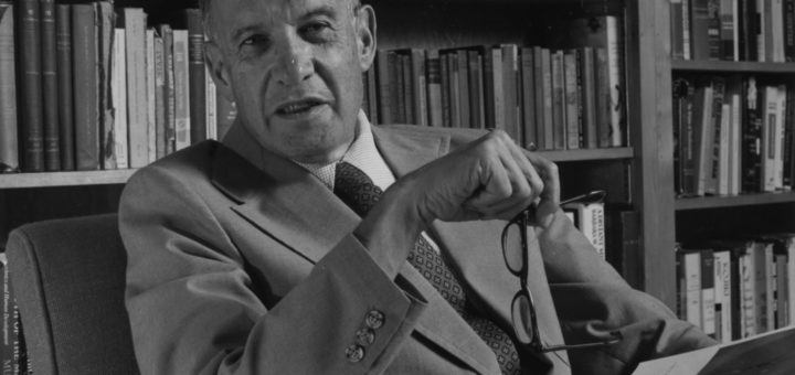 A photo of Peter Drucker at a desk in front of a shelf of books